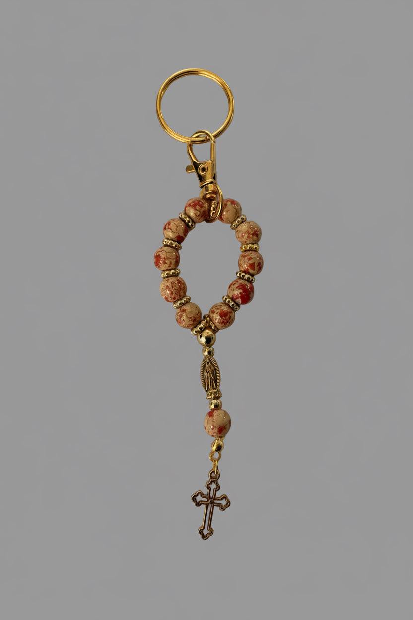 A beaded keychain featuring pinkish-red and cream stone beads with gold-toned metal spacers, a gold split ring, and a dangling gold cross charm, displayed on a textured light gray background.