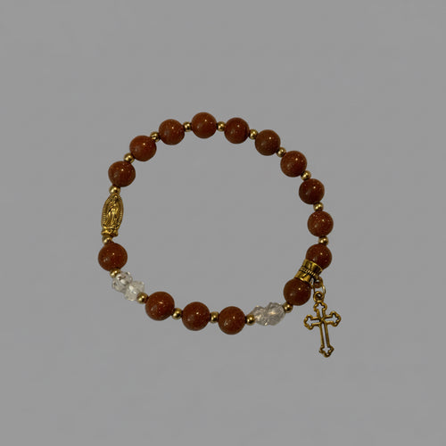 A beaded bracelet featuring round brown sandstone beads, small gold spacer beads, a gold-toned religious charm, a gold-toned cross pendant, and a cluster of clear faceted beads, displayed on a light textured surface.