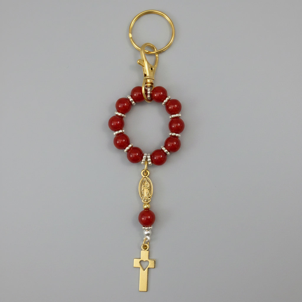 A red beaded rosary keychain with silver-tone metal accents, a split ring, and a small cross charm, shown on a textured off-white background.