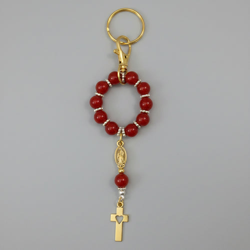 A red beaded rosary keychain with silver-tone metal accents, a split ring, and a small cross charm, shown on a textured off-white background.