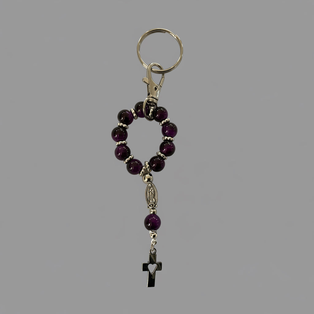 A beaded rosary keychain featuring dark purple tiger eye  beads, silver-toned metal spacers, a small silver-toned Our Lady of Fatima medal, and a black cross pendant, shown on a textured light gray background.